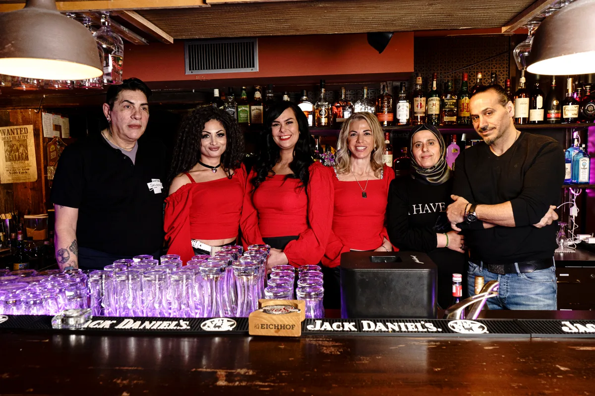 Die offizielle Eröffnung ist am 25. März. Mehrere Männer und Frauen stehen hinter einer Bartheke und schauen in die Kamera.